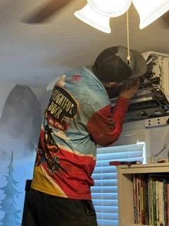 HVAC technician inspecting an indoor air conditioning unit above a bookshelf during service.